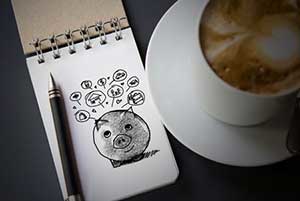 A drawing of a piggy bank with bubbles above it, on a notepad, next to a cup of coffee.