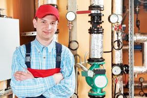 A man in work clothes stands in front of large pipes and heating equipment.