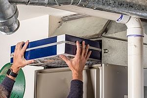 Hands are replacing a dark-blue air filter in a furnace.