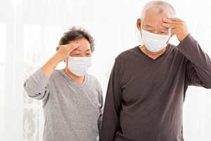 Two older adults, wearing masks, are touching their foreheads, appearing unwell.