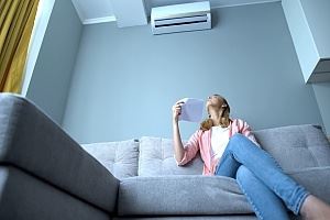A woman sits on a couch, fanning herself with paper, under an air conditioner.
