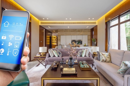 A hand holding a smart phone with home automation icons, in a spacious living room with a kitchen and sofa.