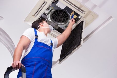 A man in work clothes is cleaning or working on a ceiling-mounted air conditioner.