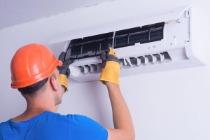 A worker in an orange hard hat and yellow gloves is working on an air conditioner unit.