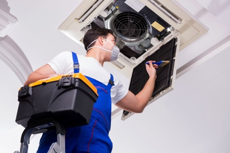 A worker in blue overalls is cleaning a ceiling air conditioner unit.