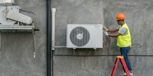 A worker in a safety vest and hard hat is on a ladder working on an air conditioning unit mounted on a gray wall.