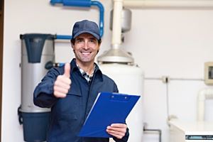 A smiling man in work clothes, giving a thumbs-up, holds a clipboard. He's in a utility room with water heater and filters.