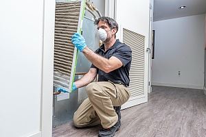 A man in work clothes and a mask is cleaning a ventilation filter.