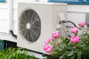 A white air conditioner unit sits outside, next to pink flowers and green plants.
