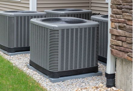 Four gray air conditioning units are on a gravel bed next to a house.