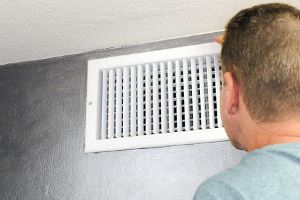 A white air vent on a gray wall. A person's head and shoulders are visible, looking at the vent.