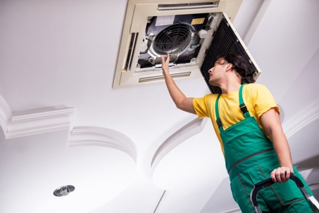 A man in green overalls is working on a ceiling-mounted air conditioner.