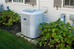 A light gray air conditioner unit sits on a stone pad outside a light beige house. Plants surround it.