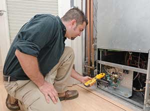 A person in light khaki pants and a dark shirt is crouched, looking at the underside of a large refrigerator unit.
