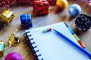 A spiral-bound notebook with colorful Christmas ornaments, small wrapped presents, and colorful letters spelling "LIST" on it. A blue pen lies beside the notebook.