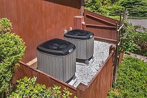 Two air conditioning units sit on a small roofed area, surrounded by a wooden fence and bushes.