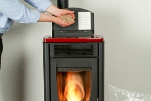 Hands are loading wood pellets into a dark gray pellet stove.