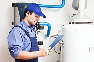A worker in blue overalls and cap is looking at paperwork near a water heater.