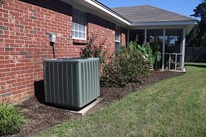 A gray air conditioner unit sits against a brick house wall, next to a screened porch and yard.