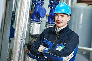 A man in a blue hard hat and work clothes stands in front of industrial machinery.