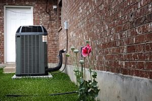 A gray air conditioner sits outside a brick wall.