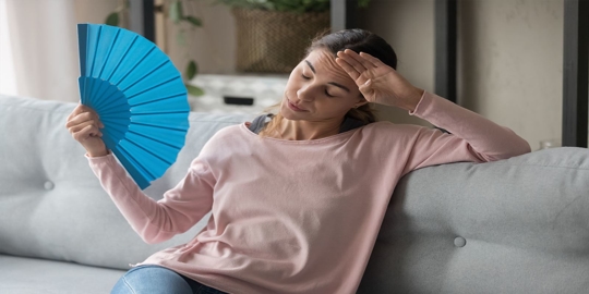A woman sits on a couch, holding a blue hand fan, looking overheated.