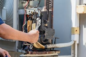 A person's hand holds a wrench near an air conditioning unit, parts of which are visible.