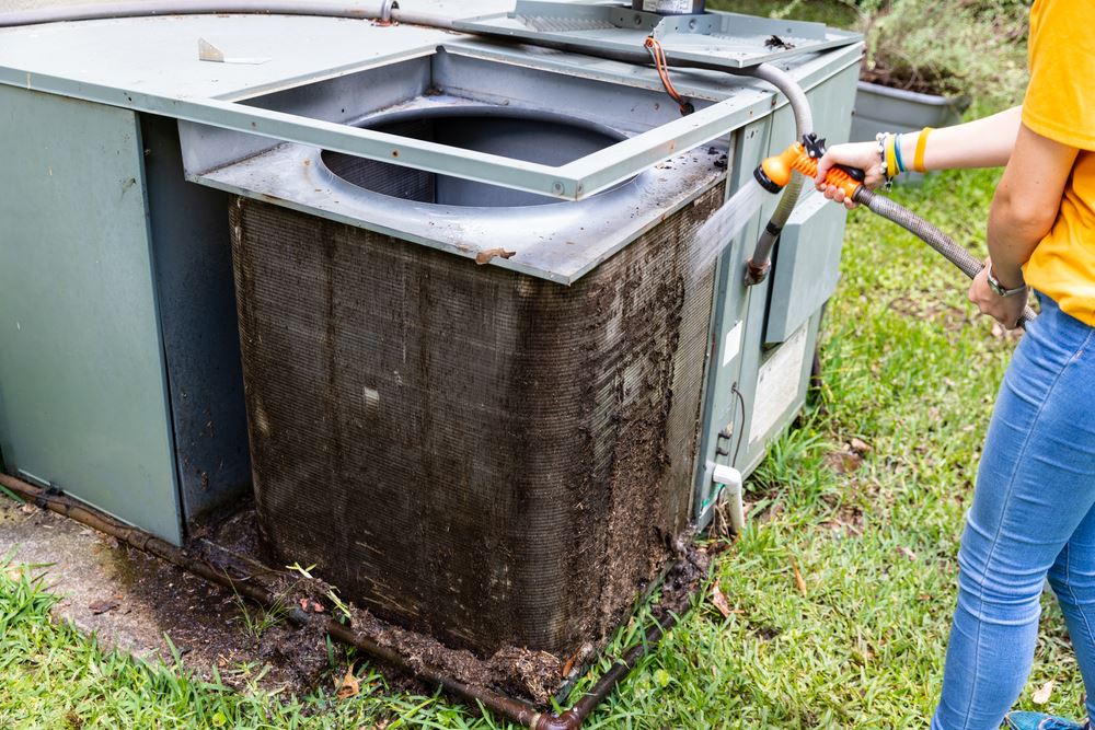 A person is cleaning a dirty outdoor air conditioner unit with a hose.