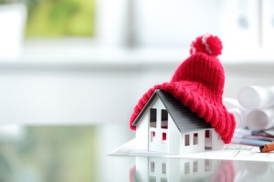 A small white house model, covered by a red knitted hat, sits on blueprints.