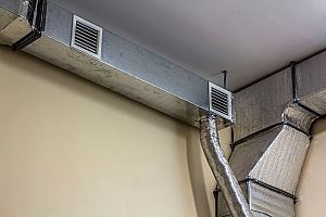 Metal air ducts and vents run along a beige wall, mounted to the ceiling.
