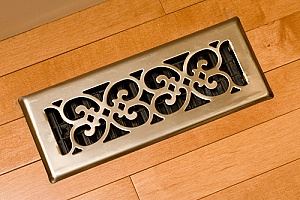 A light-colored metal floor register, rectangular and ornate, sits on light-brown wooden flooring.