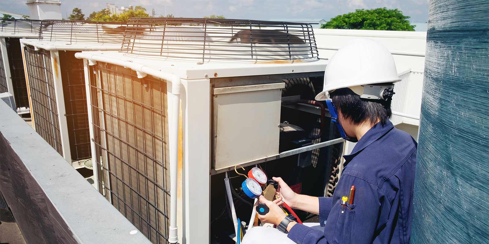 A technician in a white helmet and blue uniform is working on large air conditioning units on a rooftop.