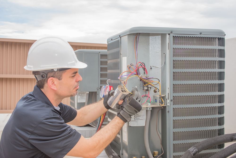 A man in a white hard hat is working on an air conditioning unit, examining and possibly repairing components.