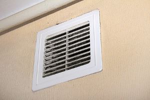 A white rectangular vent, covered in dust, is on a light beige wall.
