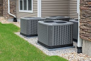 Four gray air conditioning units are outside, next to a house with a tan siding.