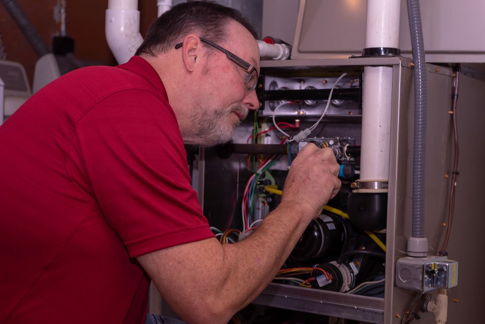 A person in a red shirt is working on a furnace, inspecting and repairing parts.