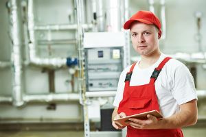 A man in red overalls and a hat, holding a tablet, stands in a mechanical room with pipes and equipment.
