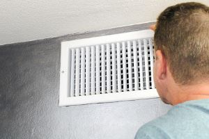 A white air vent on a gray wall. A person's head and shoulders are visible, looking at the vent.