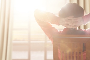 A person sits in a chair, hands behind head, relaxing by a sunny window.