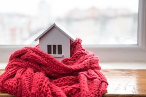 A small white house figurine is wrapped in a red knitted scarf, near a window.