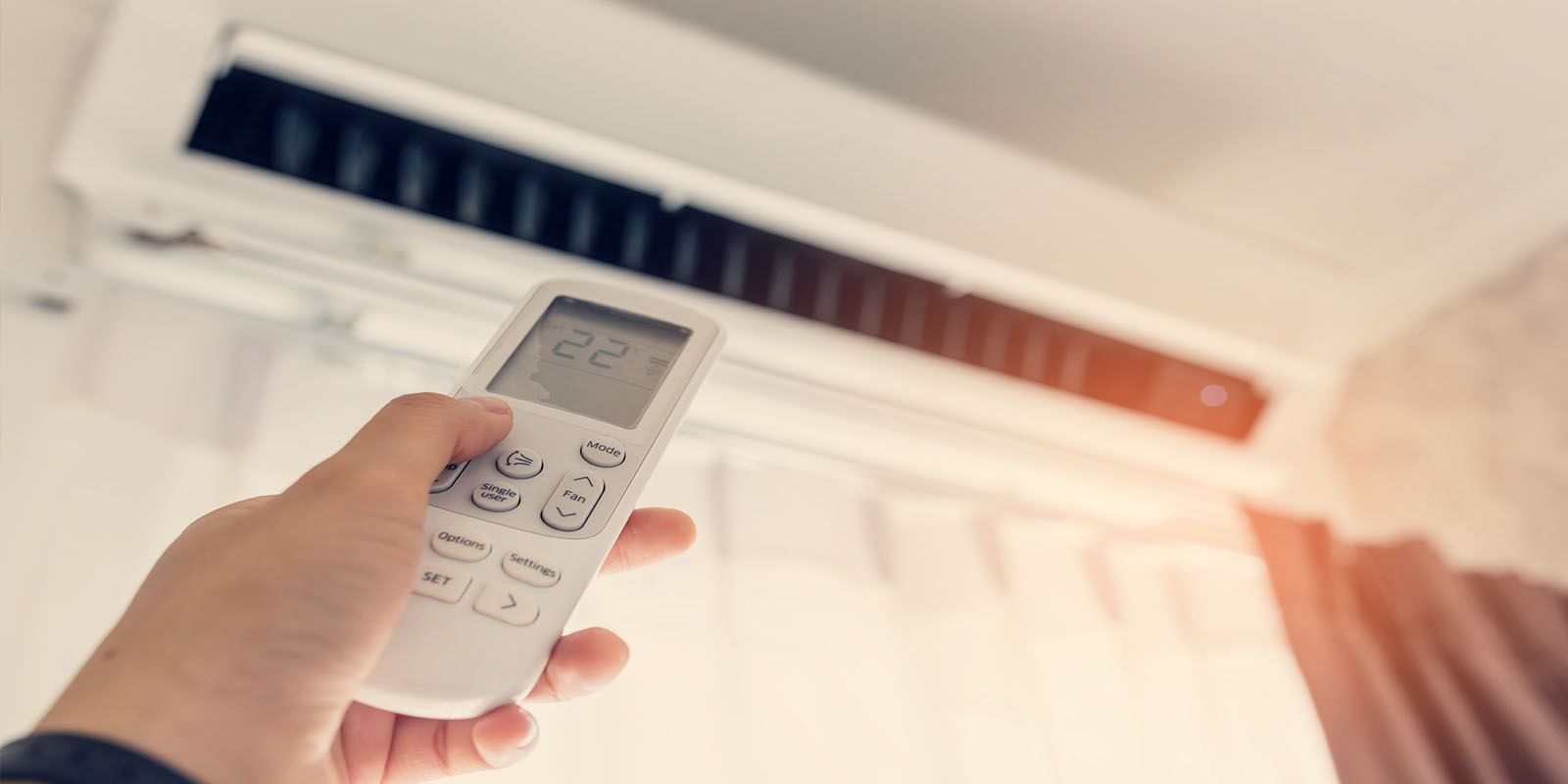 A hand holds an air conditioner remote, with the unit above.