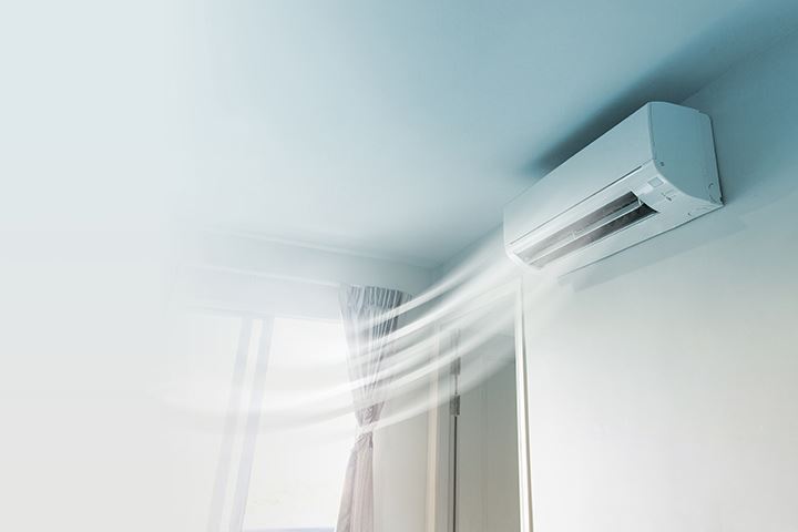 A white air conditioner is mounted on a wall, blowing air towards a window.
