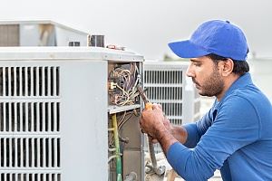 A man in a blue cap and shirt is working on an air conditioning unit outdoors.
